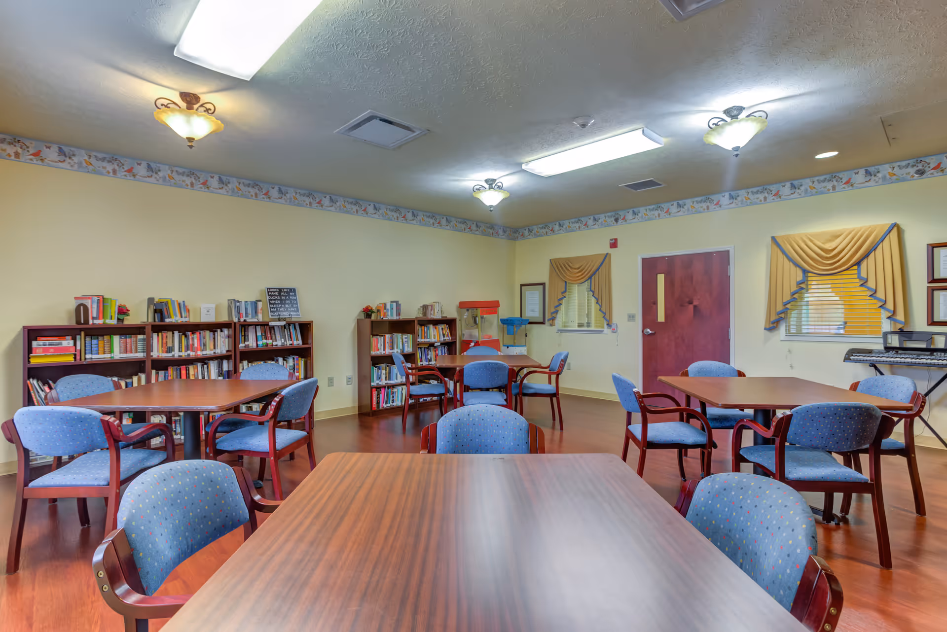 A senior living facility common area with tables, blue chairs, and bookshelves.