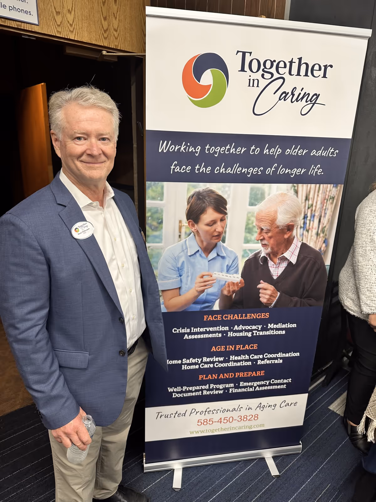 Man in blue suit stands beside Together in Caring banner inside a facility.
