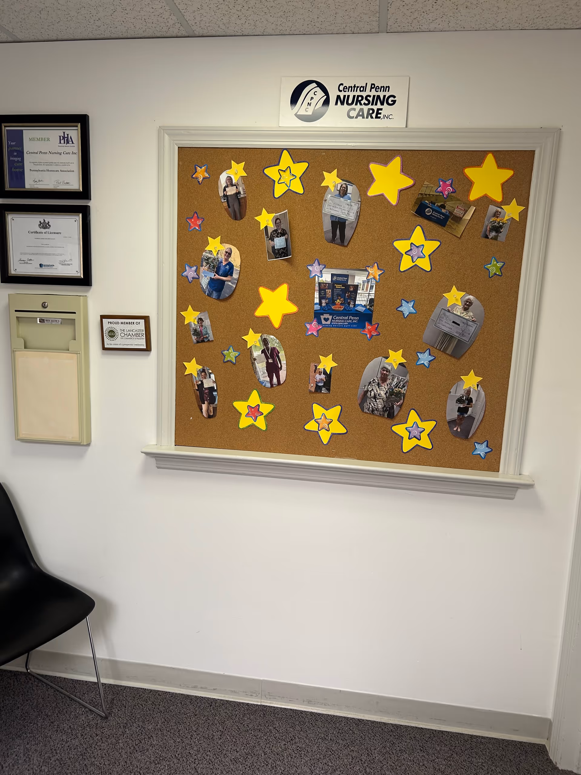 Interior hallway with a corkboard displaying photos and yellow star shapes, framed certificates, and a chair.