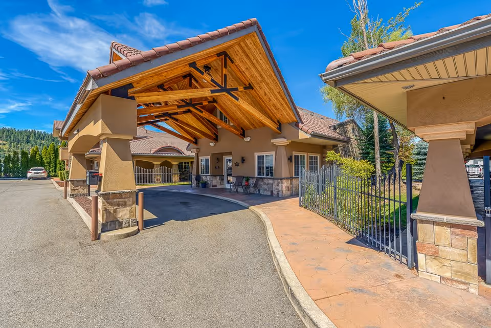 Entrance of Pinehurst Senior Community featuring a covered driveway with wooden beams and stone pillars, adjacent to a paved walkway and landscaped greenery under a bright blue sky.