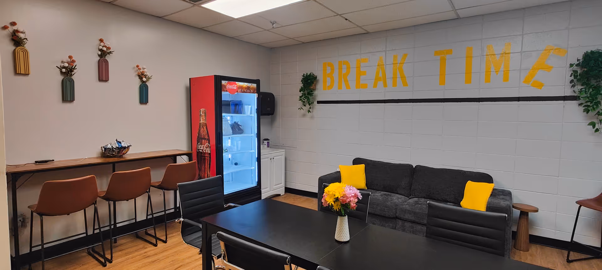 A break room with a black couch adorned with two yellow pillows, a black table with black chairs, and a vase of flowers on the table. On the wall behind the couch, the words 'BREAK TIME' are painted in yellow. To the left, there is a Coca-Cola vending fridge, a counter with three brown chairs, and decorative wall hangings with flowers. The room has wooden flooring and white walls with some green plants on the wall.