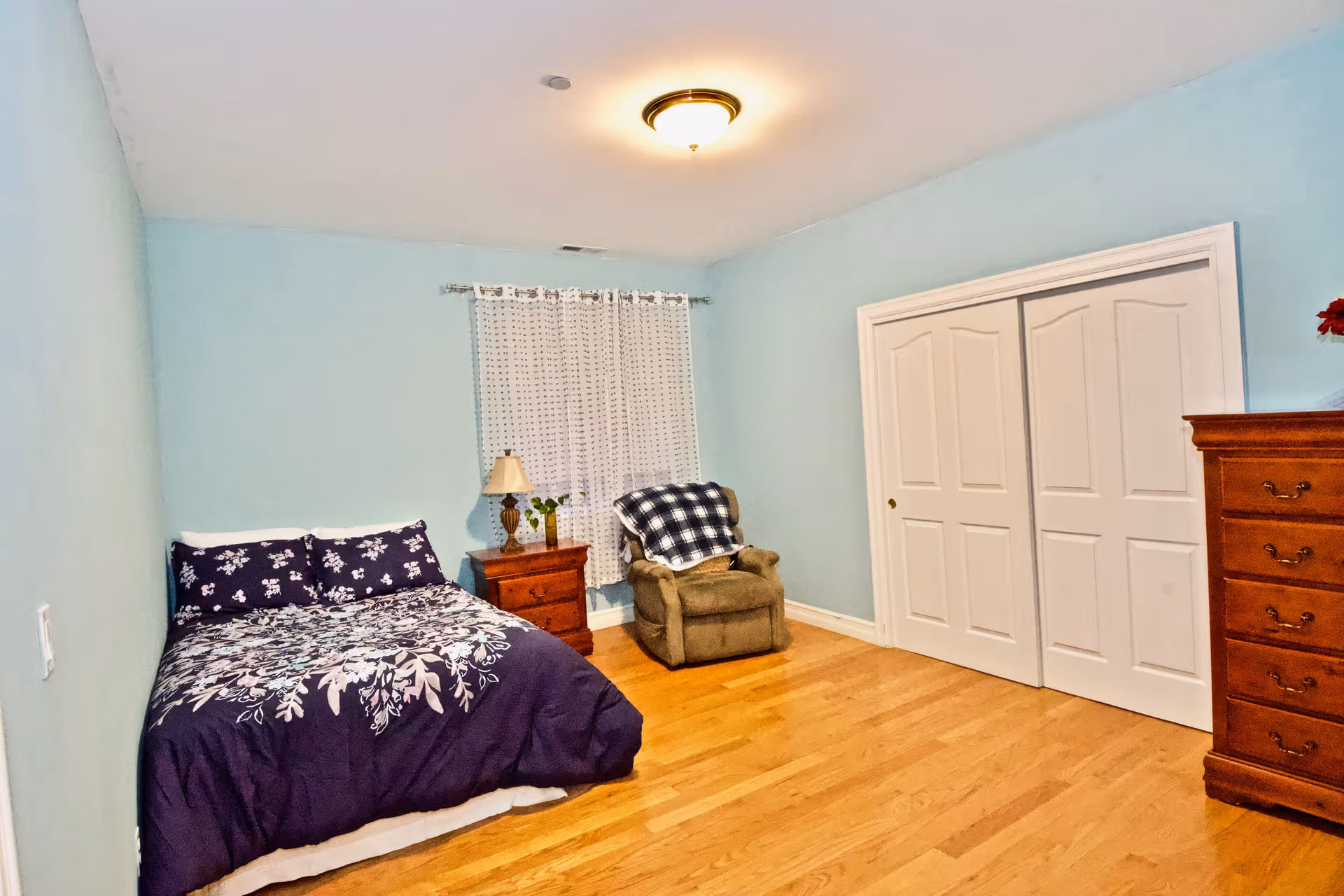 Light blue bedroom with a bed, nightstand, dresser, chair, and double closet doors.