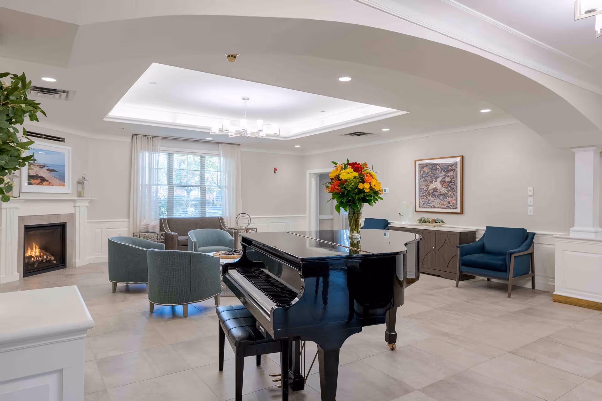 Bright senior living lounge featuring a grand piano, seating area, fireplace, and floral centerpiece.