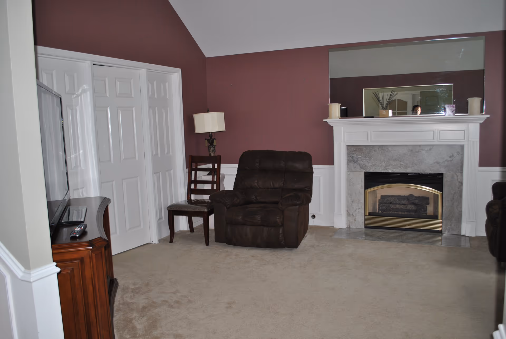 Carpeted living room with a brown recliner and wooden chair facing a fireplace with a large mirror and a TV on a wooden stand.