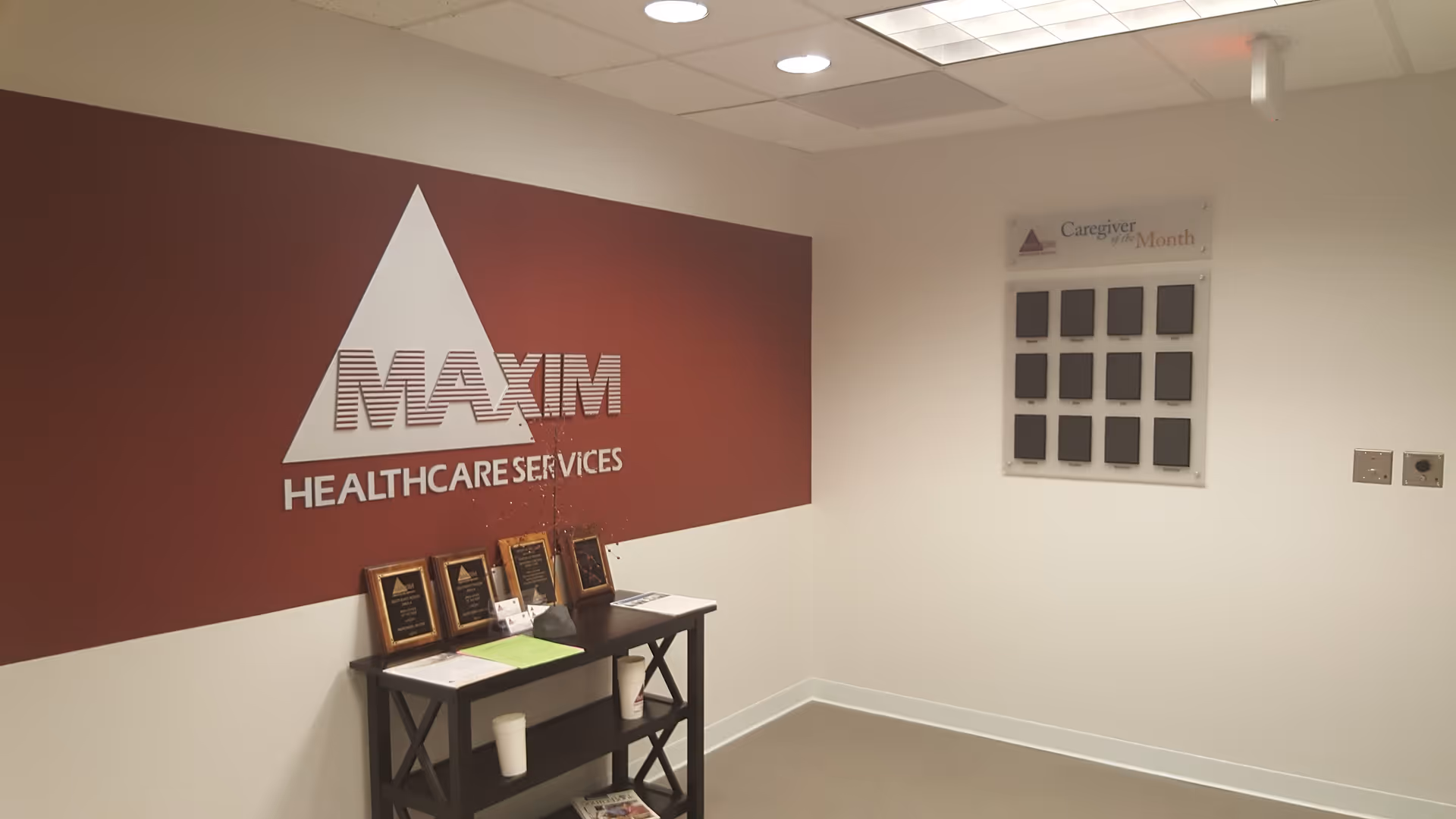 Interior reception area with Maxim Healthcare Services logo on burgundy wall and an awards table.