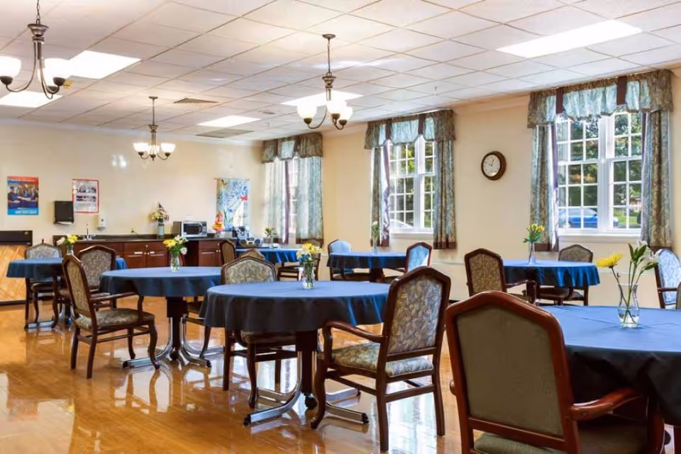 Bright communal dining room with round tables covered in blue tablecloths, upholstered chairs, chandeliers, and windows with curtains.