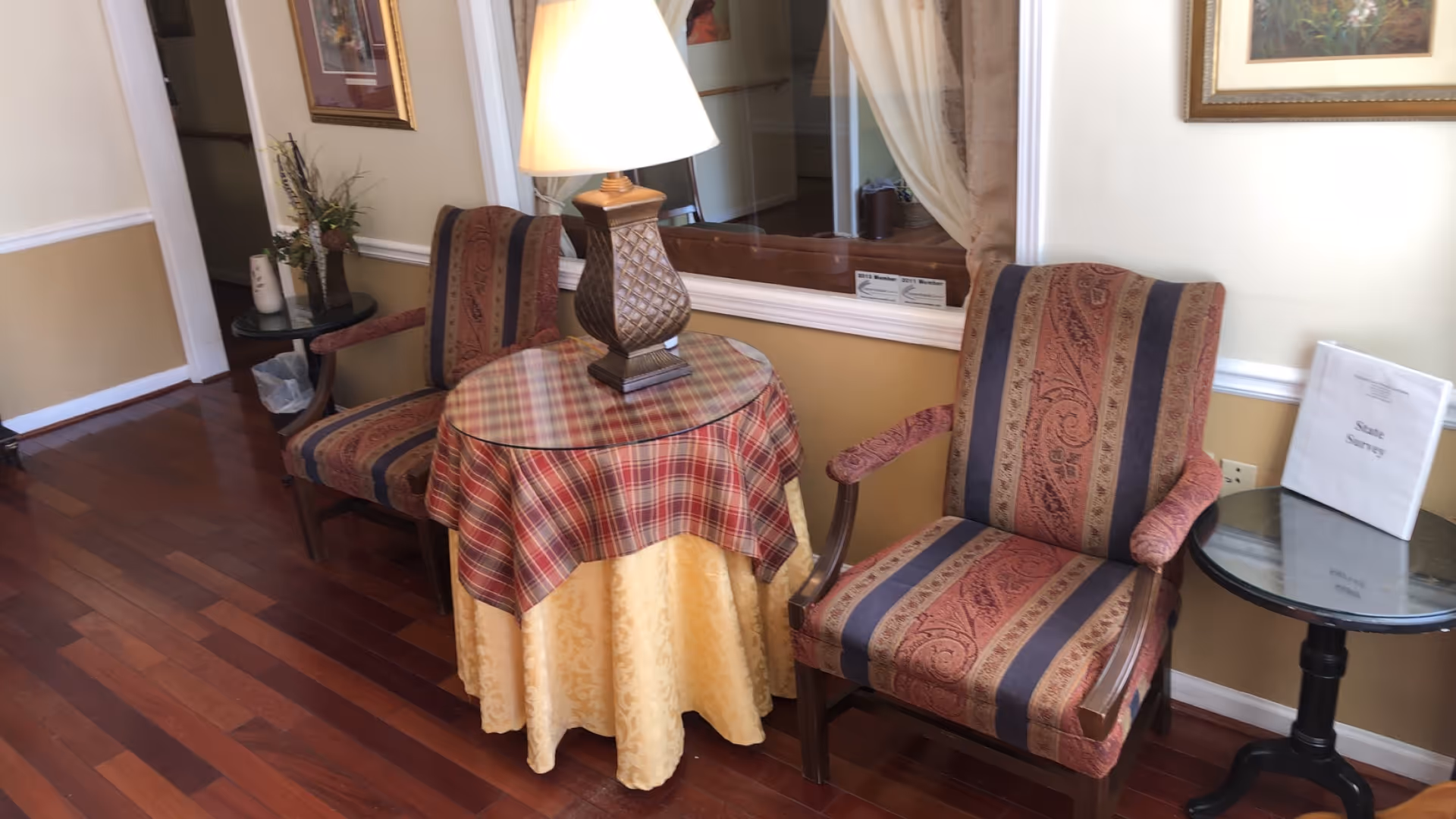 A cozy sitting area with two upholstered armchairs featuring a red and blue patterned fabric. Between the chairs is a round table covered with a yellow and red plaid tablecloth, topped with a decorative lamp. Another small round table with a glass top holds a binder labeled 'State Survey'. The room has wooden flooring and light-colored walls with white trim.