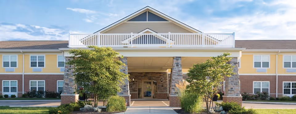 Front exterior view of a senior living facility named Traditions at Reagan Park, featuring a covered entrance supported by stone pillars, landscaped greenery, and a two-story building with yellow and brick facade under a partly cloudy sky.