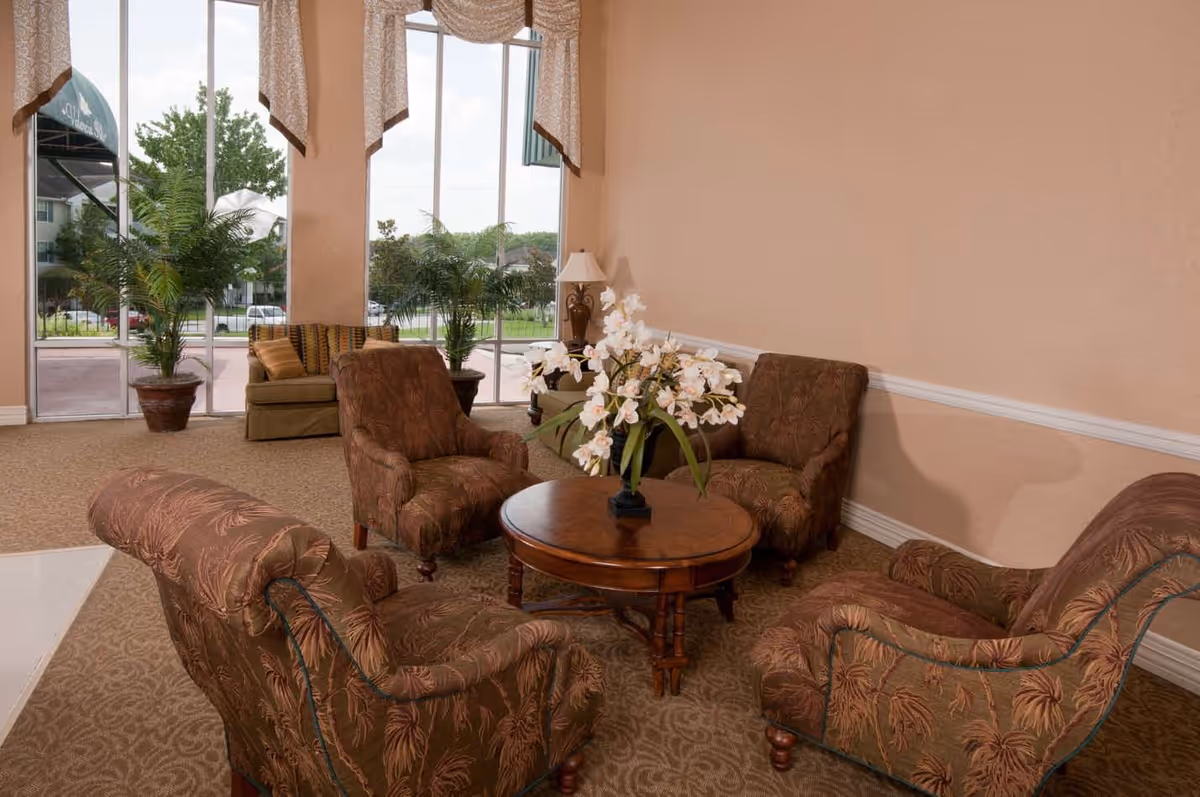 A cozy seating area with four upholstered armchairs arranged around a round wooden table with a floral centerpiece. The room has large windows with beige curtains, potted plants near the windows, and a beige carpeted floor.