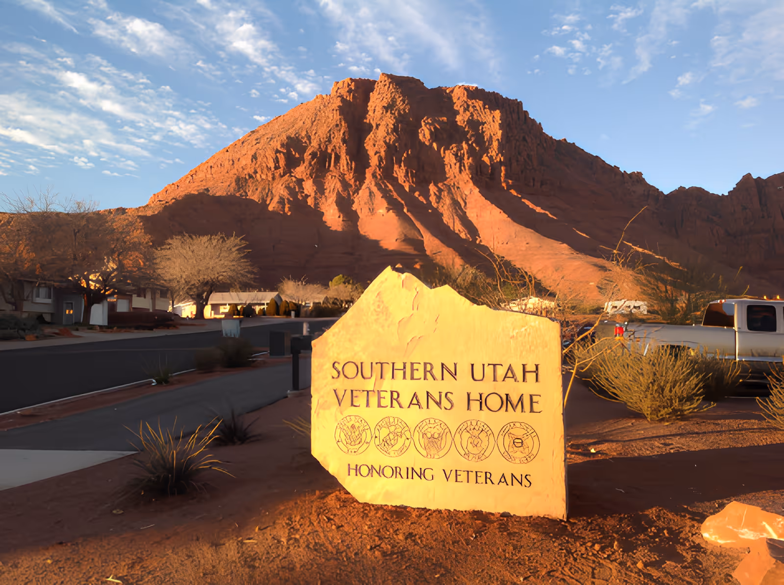 A large stone sign at the entrance of Southern Utah Veterans Home with the text 'Southern Utah Veterans Home Honoring Veterans' and military service emblems. The sign is set against a backdrop of a red rocky mountain and a clear blue sky, with some desert vegetation and a pickup truck visible nearby.