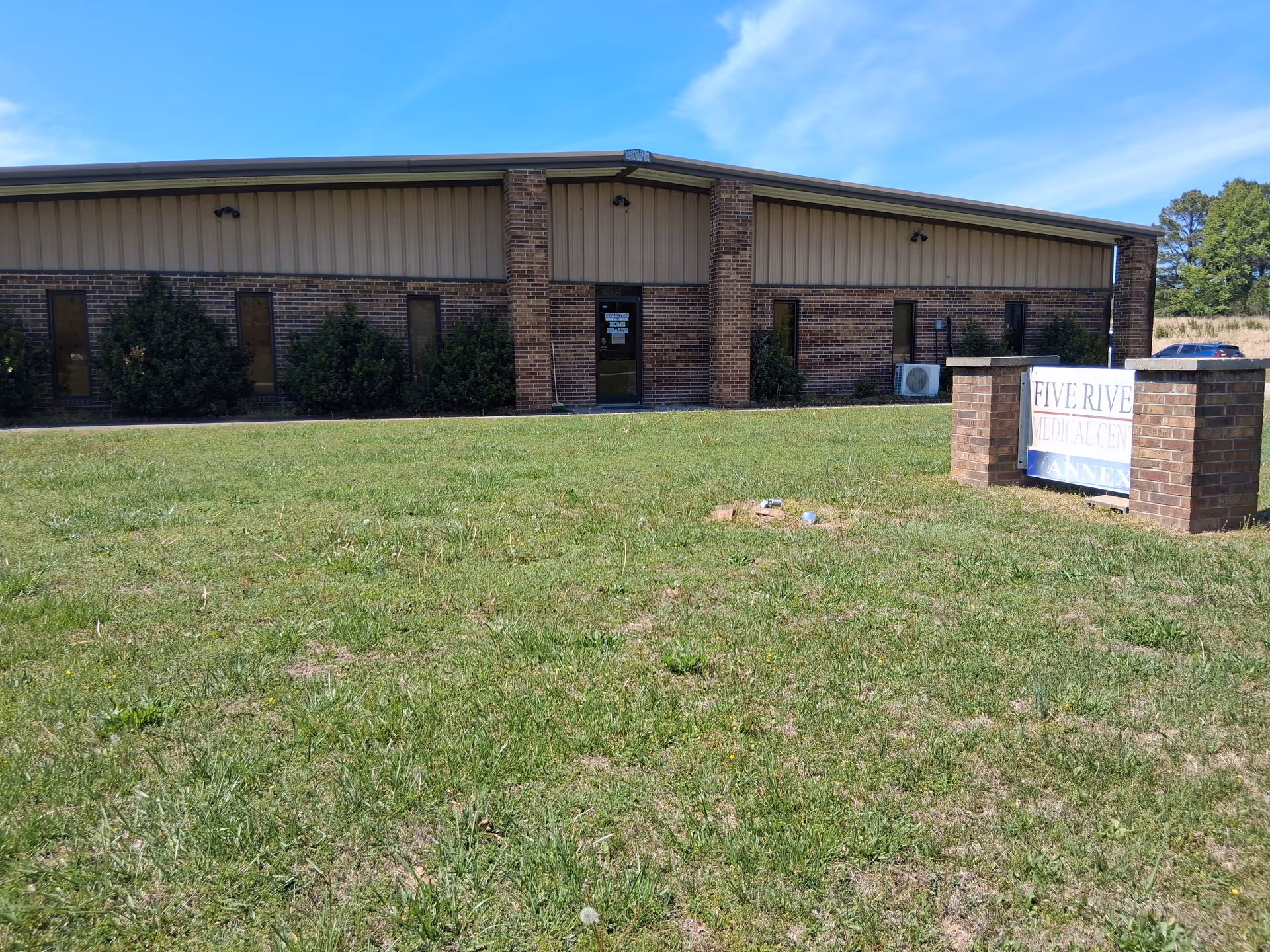 Front of a brick medical facility with a sign and grassy foreground.