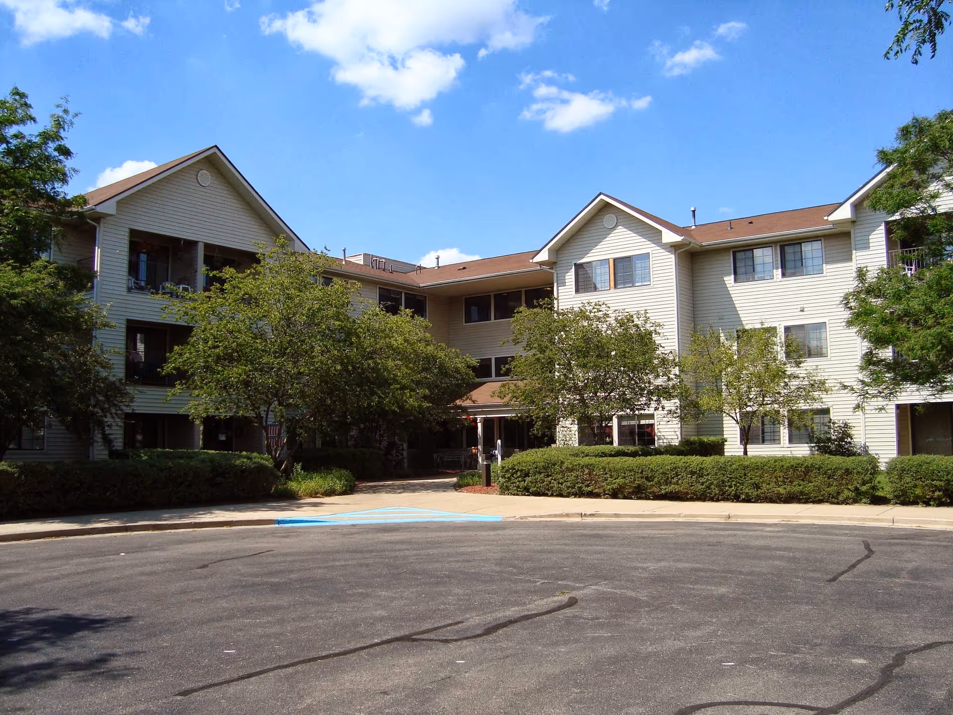 Three-story beige senior living building with trees, shrubs, and a driveway under a blue sky.