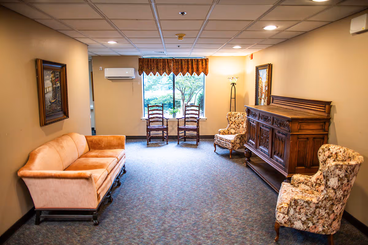 A cozy sitting area with a peach sofa, floral armchairs, a carved wooden cabinet and two wooden chairs by a window.