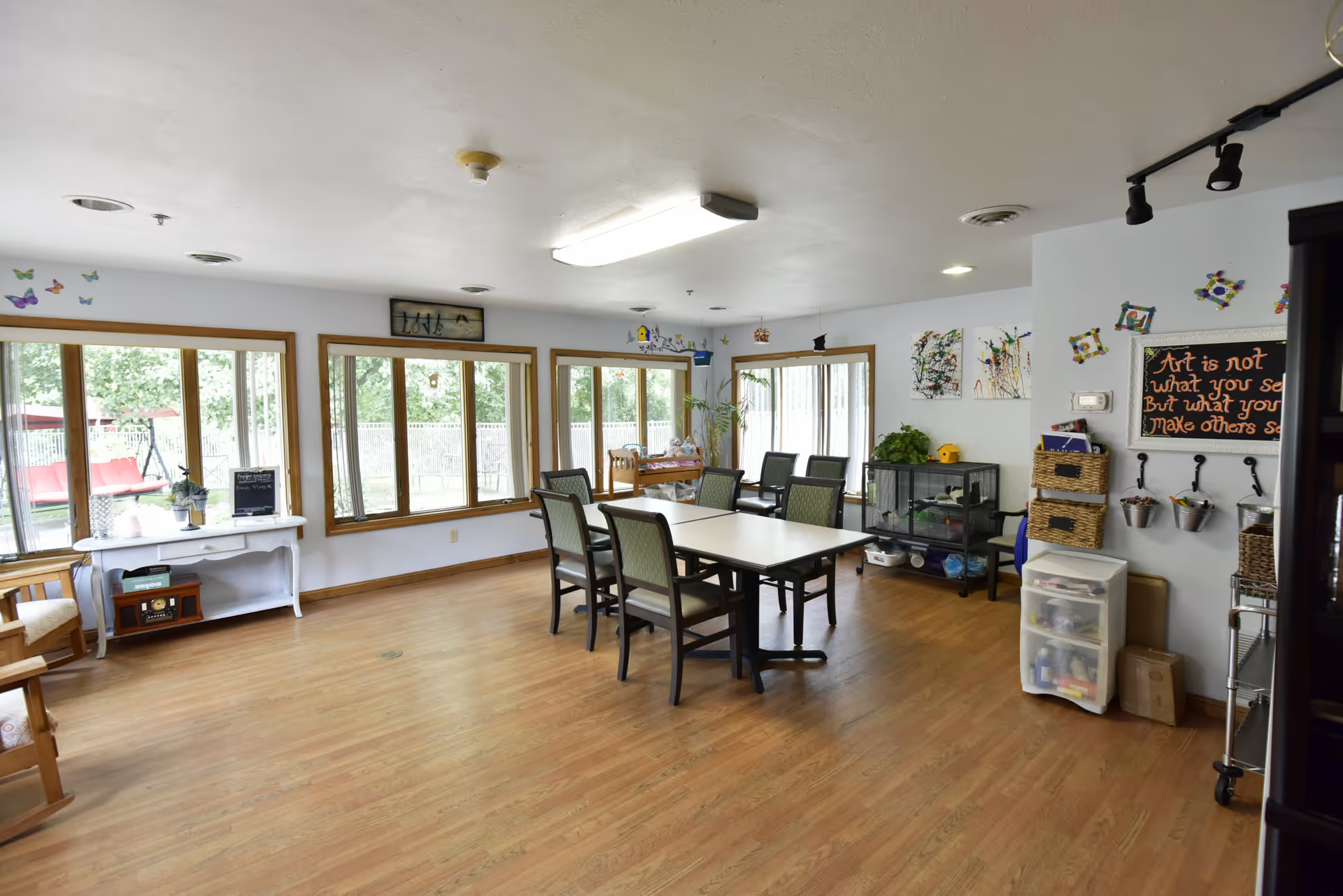 A bright, spacious room with large windows letting in natural light. The room has wooden floors and several chairs arranged around two tables in the center. There are various decorations on the walls, including colorful artwork and a chalkboard with a quote. A small white table with a plant and a radio sits near the windows, and a cage with plants and supplies is against the far wall. The room appears to be a communal or activity area in a senior living facility.
