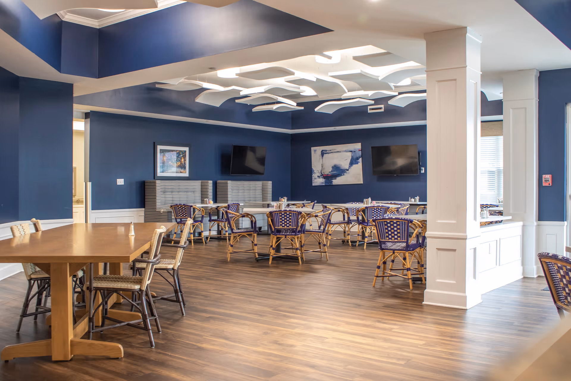 A spacious dining room with wooden floors, blue walls, and modern white ceiling fixtures. The room contains multiple tables with wicker chairs featuring blue cushions. Two flat-screen TVs are mounted on the walls, and there are framed artworks displayed. The room is well-lit with natural light coming through windows.