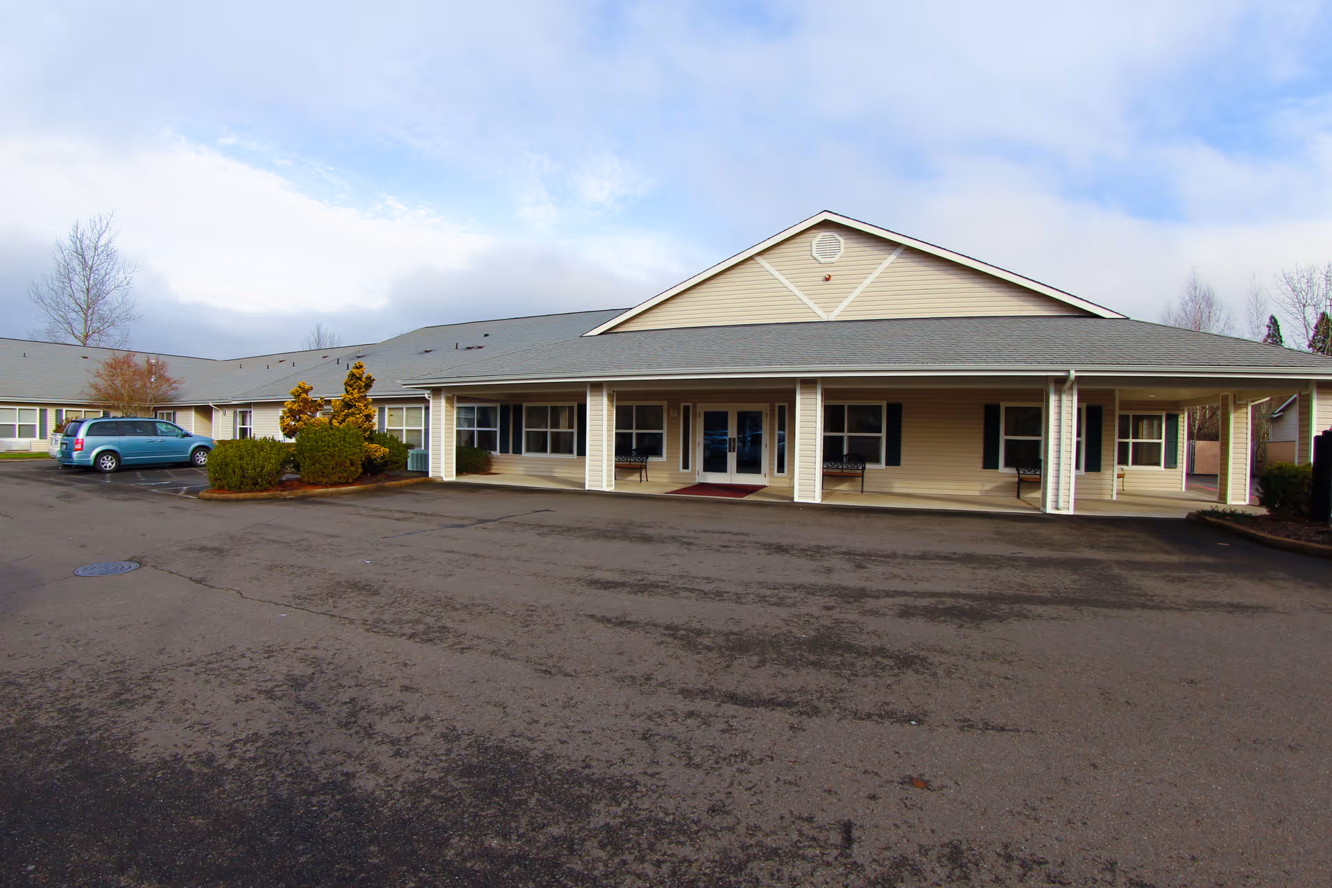 Front exterior of a single-story beige building with a covered entrance, small landscaping, and an asphalt parking lot with a blue minivan at left.