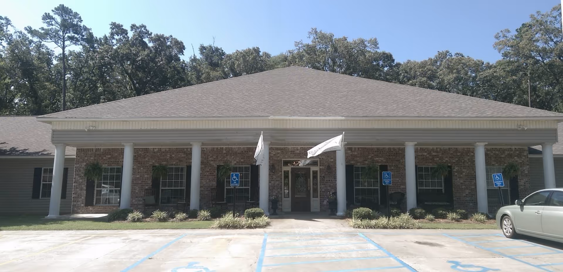 Front exterior view of a single-story brick building with a covered entrance supported by white columns. There are two flags flying above the entrance and several handicap parking spaces in front. The building is surrounded by trees and greenery.