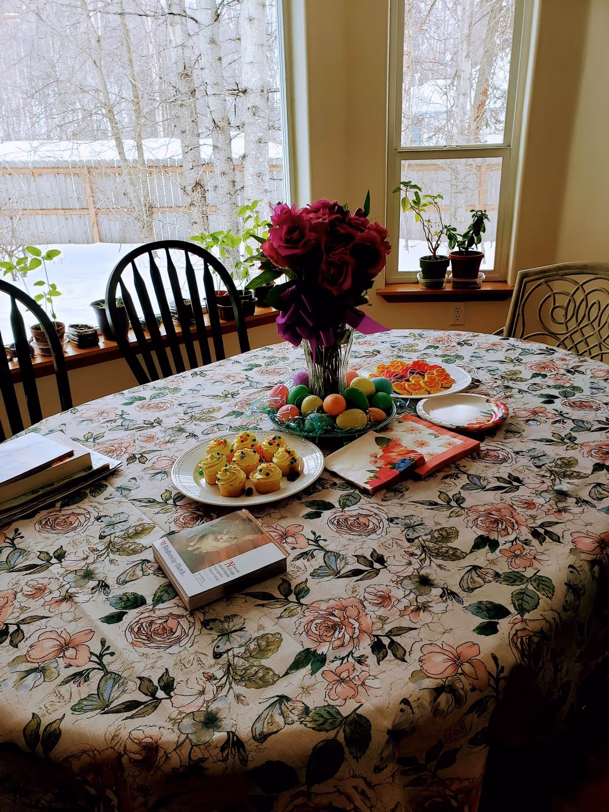 Round dining table with floral tablecloth, centerpiece flowers, cupcakes, and colorful Easter eggs indoors.