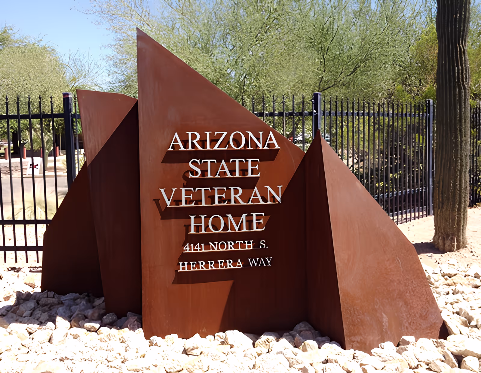 Rust-colored metal entrance sign reading "Arizona State Veteran Home" set among rocks with a black fence, cactus, and desert trees behind it.