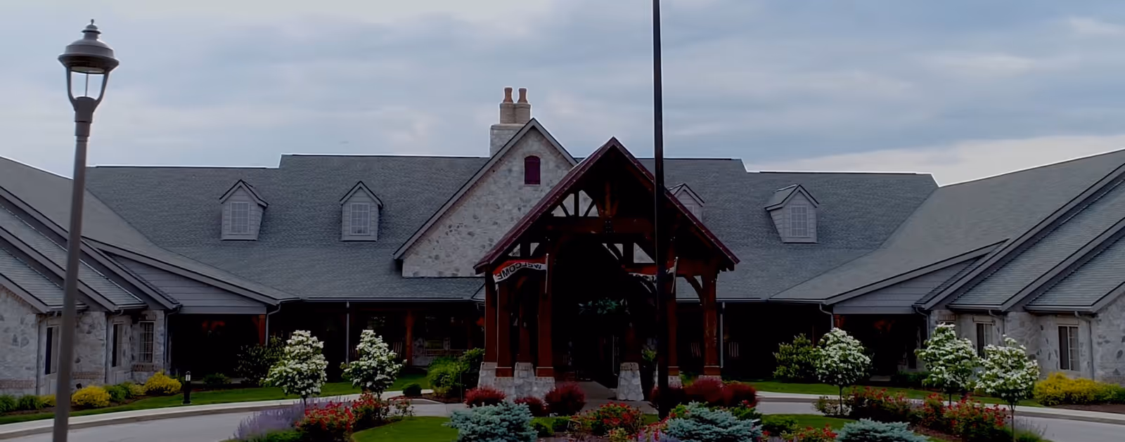 Stone-faced senior living facility building with a covered wooden entrance, circular driveway and landscaped flowerbeds in front.