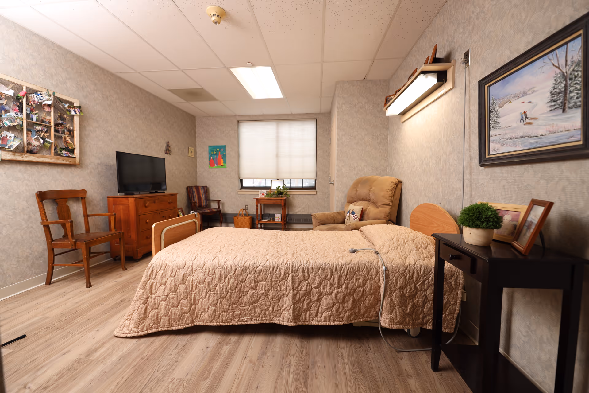 A cozy assisted-living bedroom with a bed, recliner, TV, chairs, and nightstand in a softly lit room.