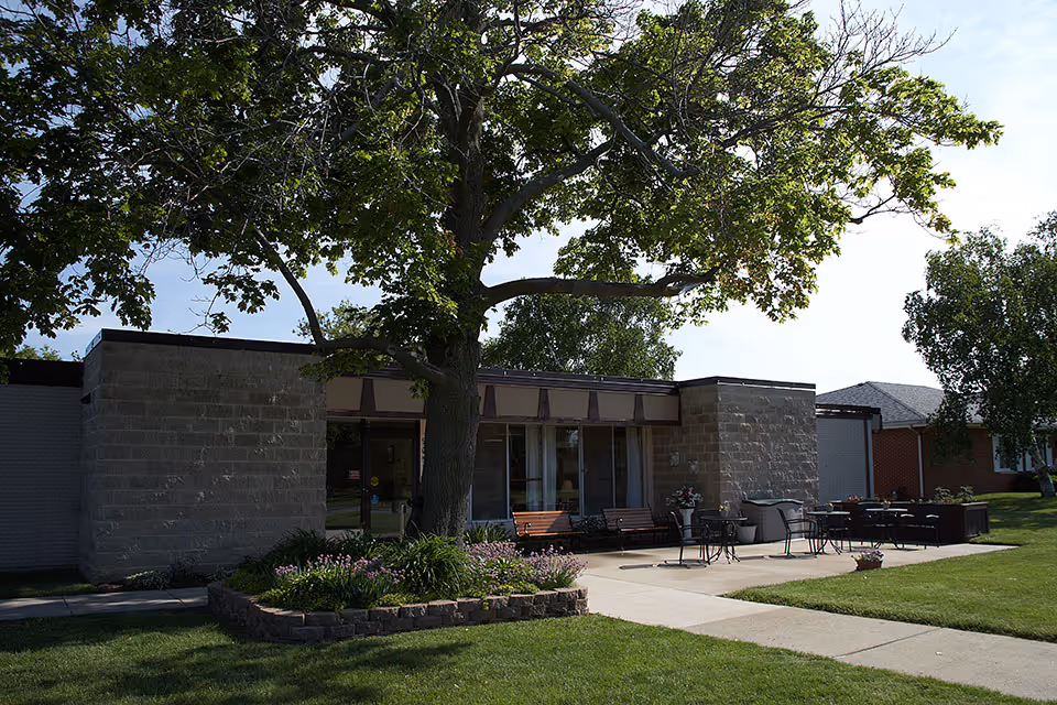 Outdoor patio area of a senior living facility with a large tree providing shade. The patio has several benches, tables, and chairs arranged on a concrete surface. The building is made of light-colored bricks with large windows and a door visible. There is a well-maintained lawn and flower beds surrounding the patio.