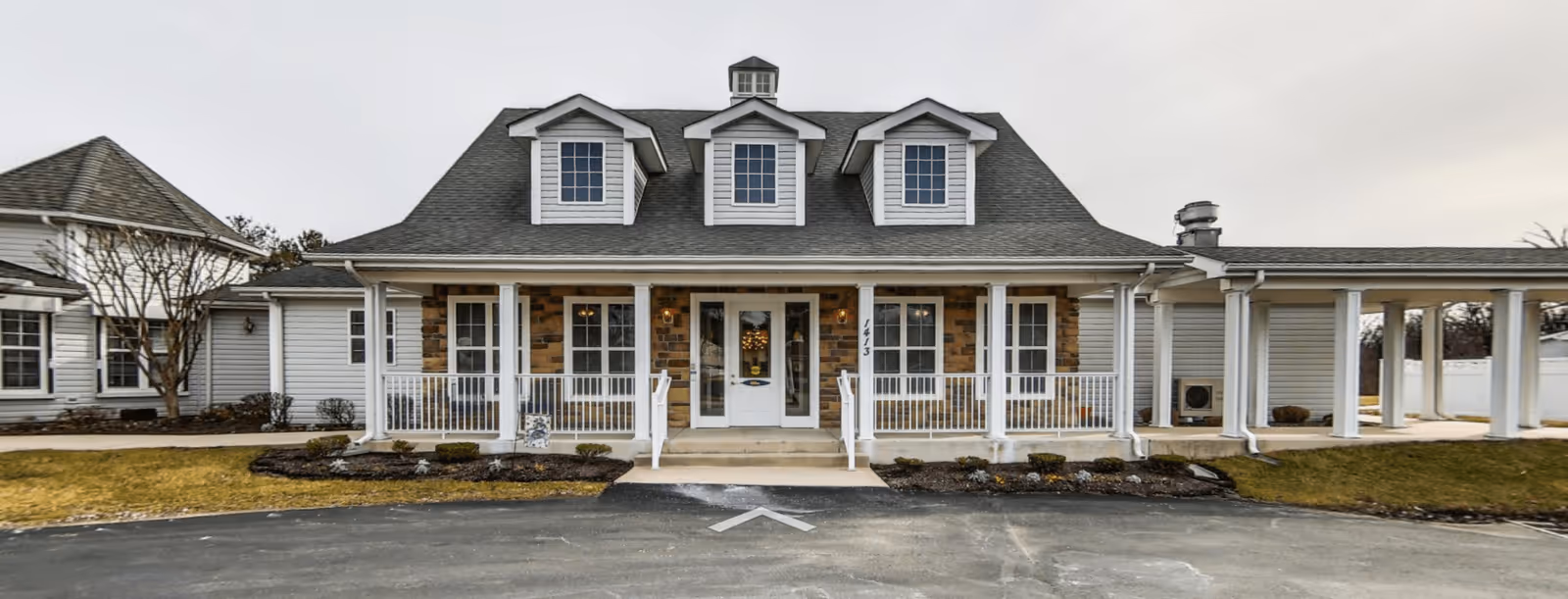 Front exterior of a single-story senior living building with a covered porch, white columns, dormer windows, and a driveway.