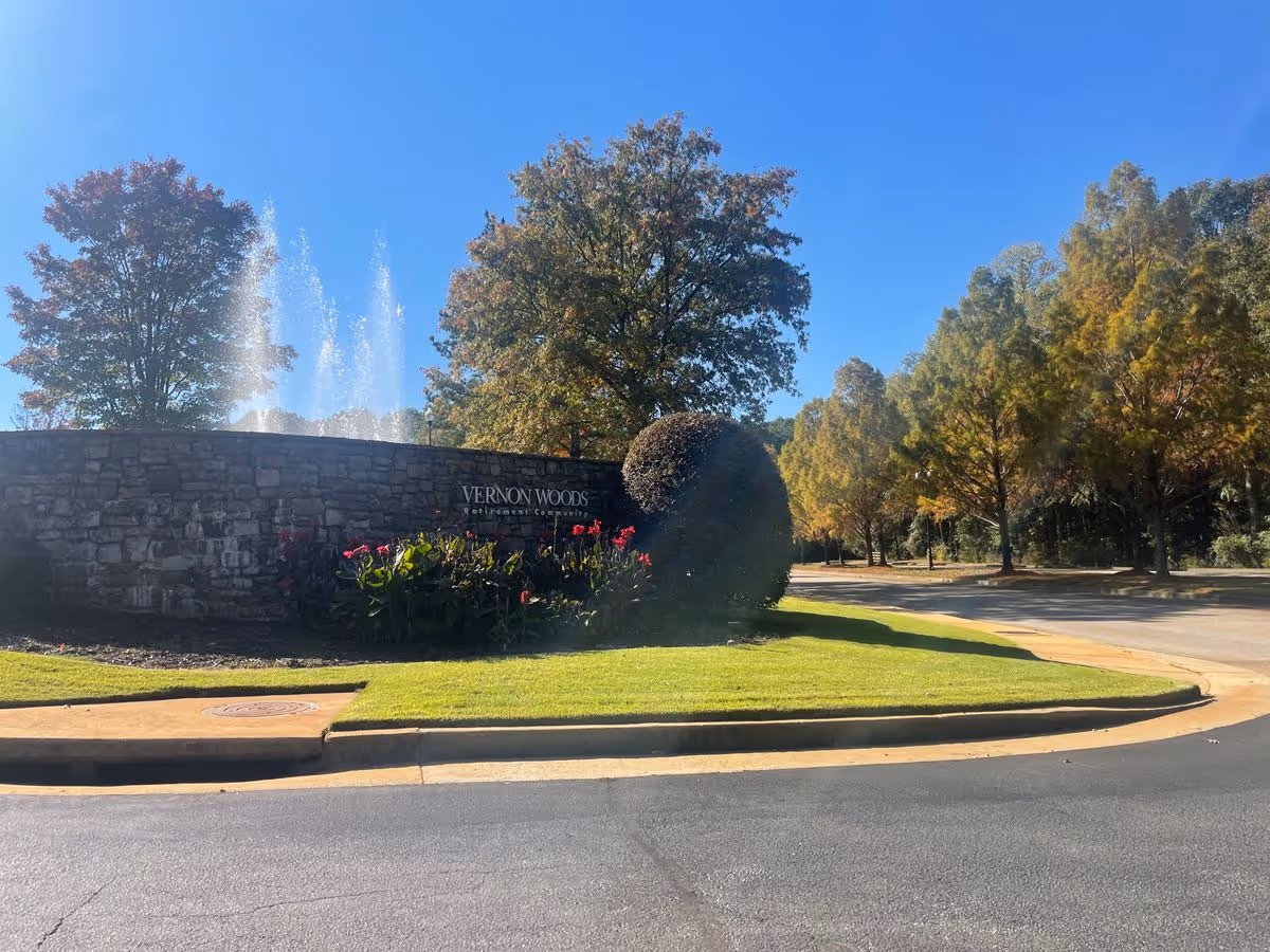 Stone entrance wall reading 'Vernon Woods' with fountains, landscaped flowers, and trees under a clear blue sky.