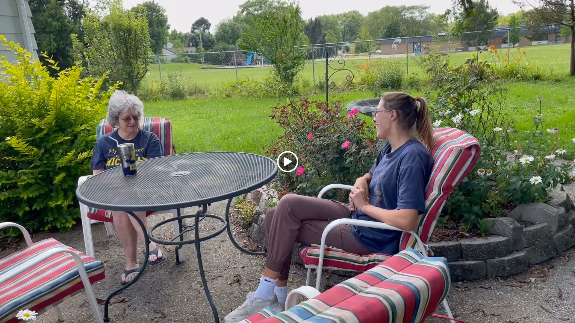 Two women sitting and talking outdoors on striped cushioned patio chairs around a round metal table, with green grass, bushes, and flowers in the background.