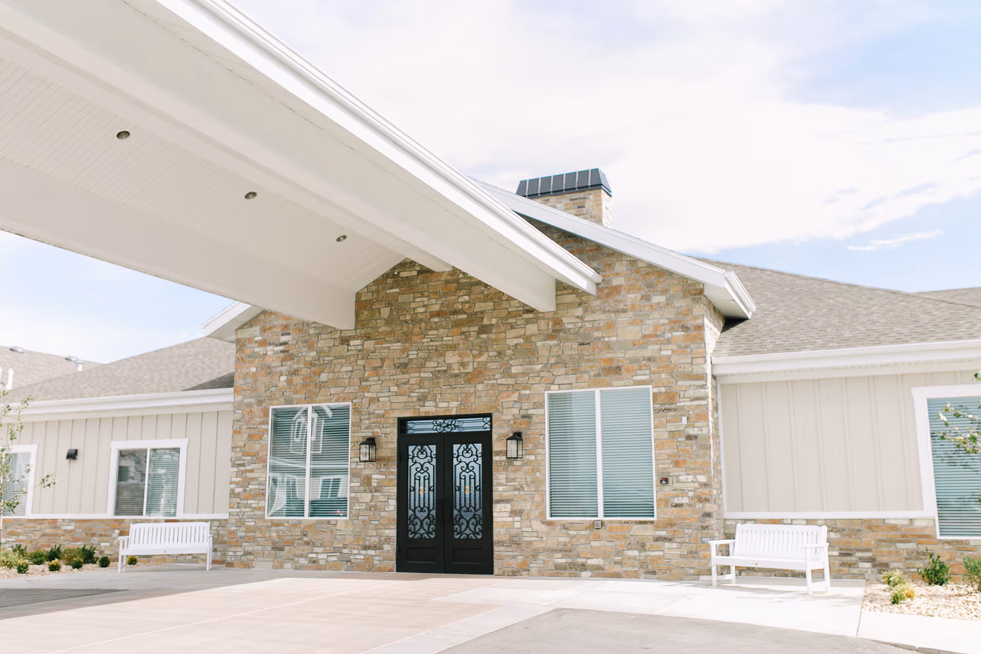 Front entrance of Whisper Cove Assisted Living and Memory Care with a stone façade, decorative double black doors, a covered portico, and white benches.
