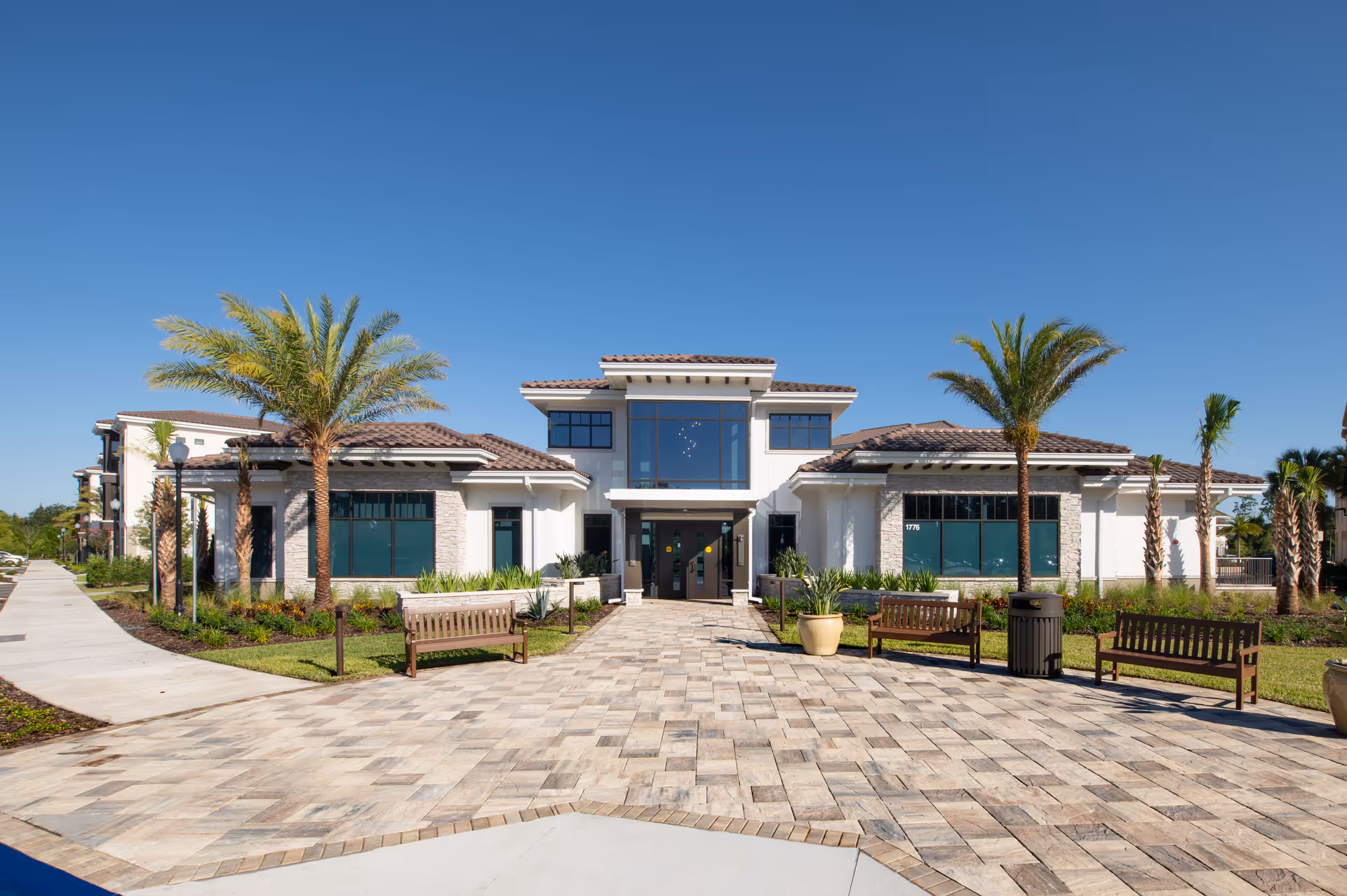 Front exterior view of The Mayflower at Winter Park facility with a paved walkway leading to the entrance, surrounded by palm trees, benches, and landscaped greenery under a clear blue sky.