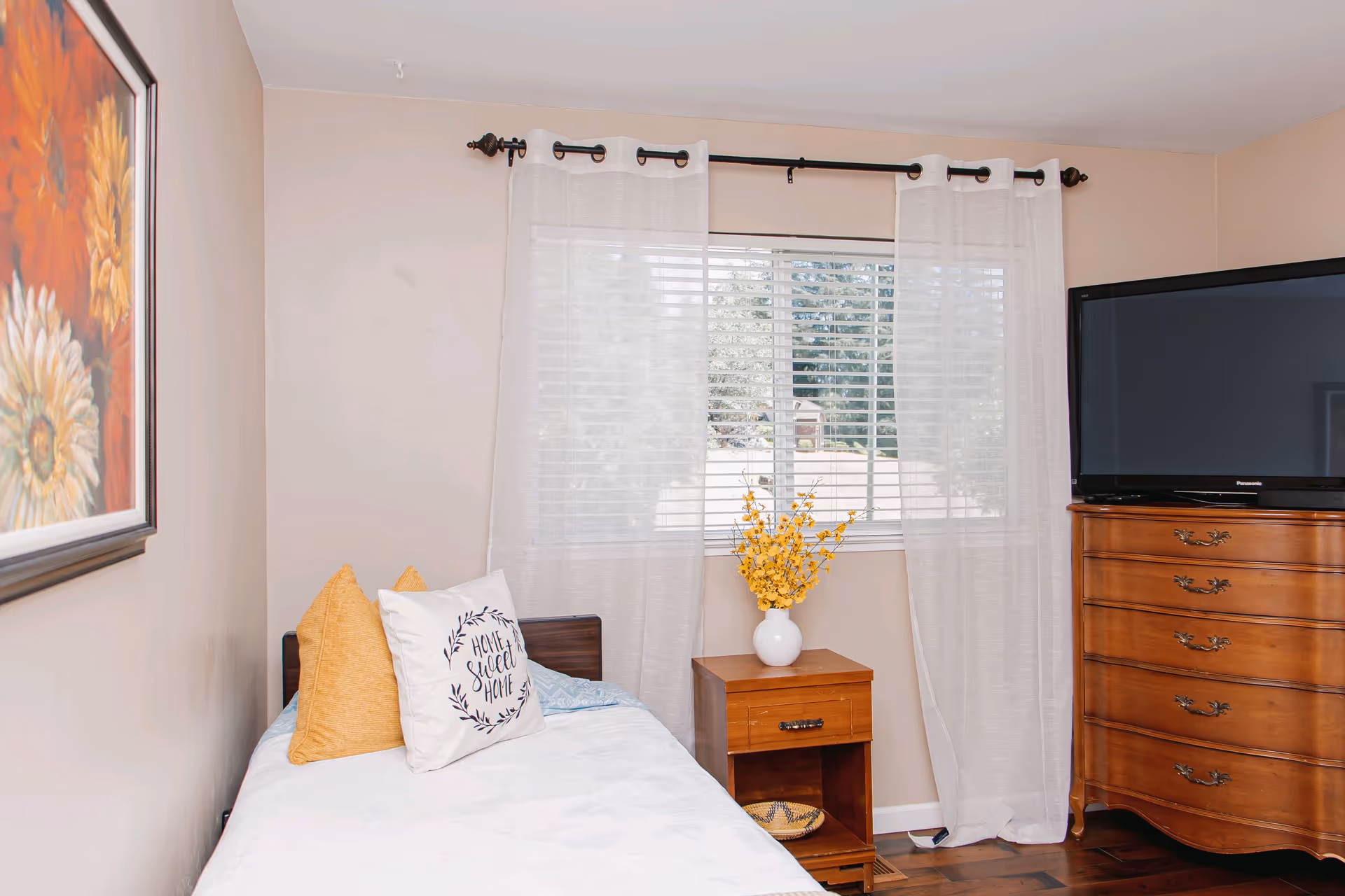Bedroom with bed, nightstand, dresser with TV, and window with sheer curtains.