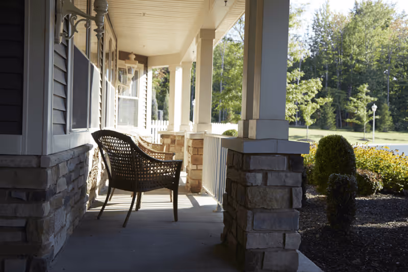 Covered front porch with a wicker chair between stone columns overlooking a landscaped yard.