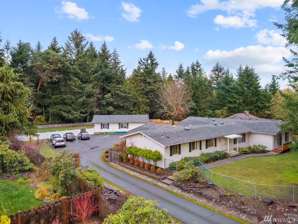 Aerial view of Harbor Heritage Adult Family Home, a single-story building with a gray roof surrounded by green trees and landscaped gardens. There is a driveway leading to a parking area with several cars parked. The sky is partly cloudy with blue patches.