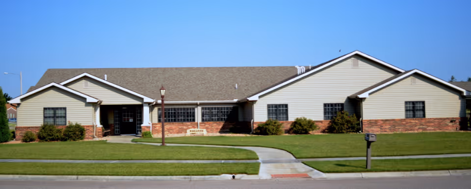 Single-story brick-and-siding senior living building with a central entrance, lawn, and curved walkway under a clear blue sky.