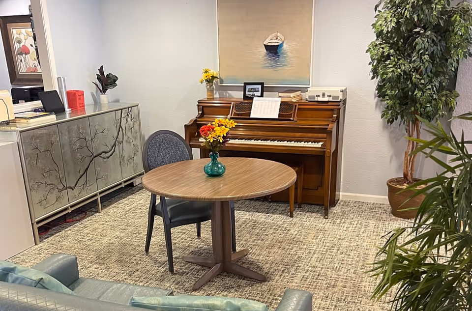 Lounge area with round table, piano, glass-front cabinet, and potted plants in a senior facility.