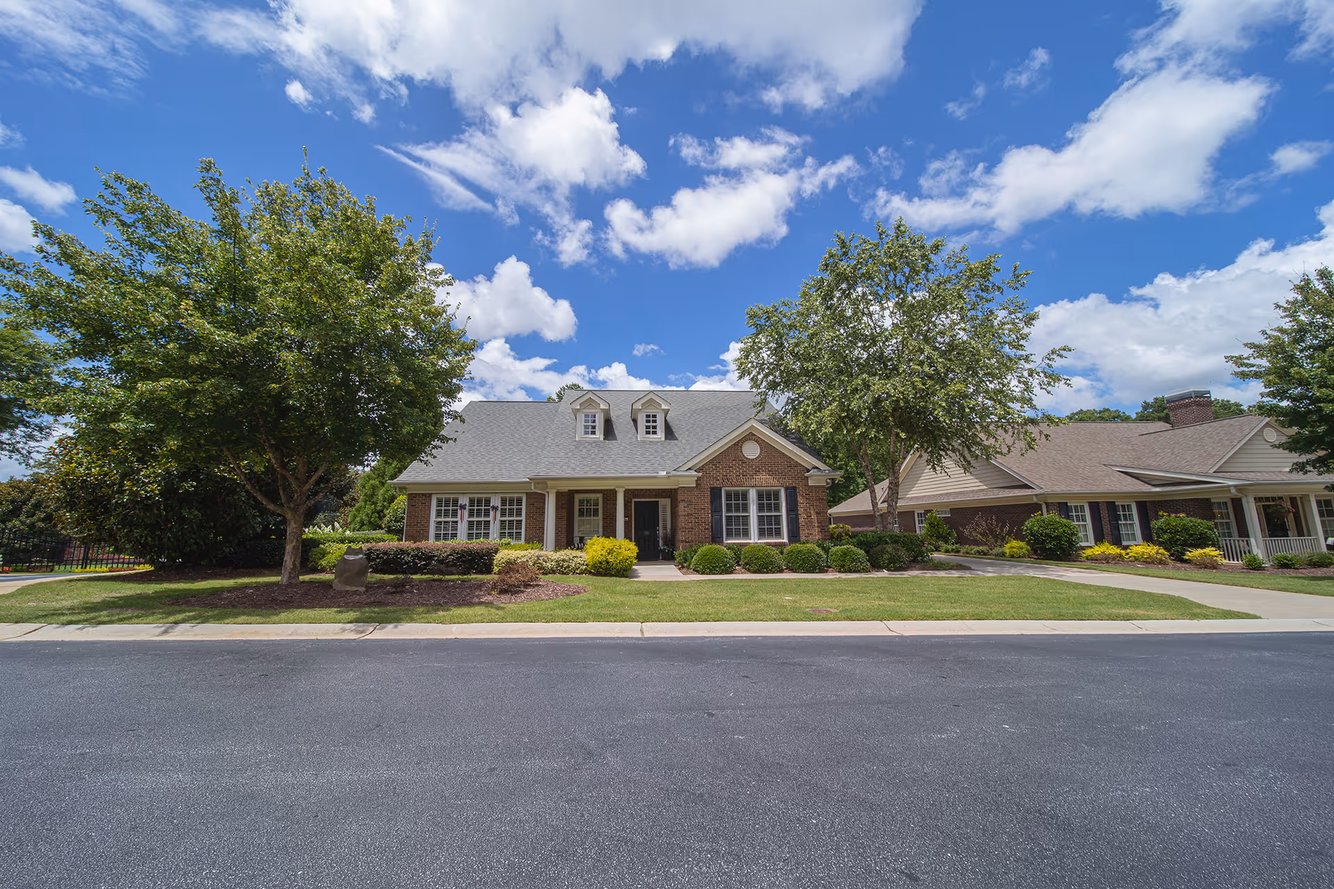 Exterior view of a single-story brick building with a gray roof, surrounded by green trees and shrubs under a partly cloudy blue sky.