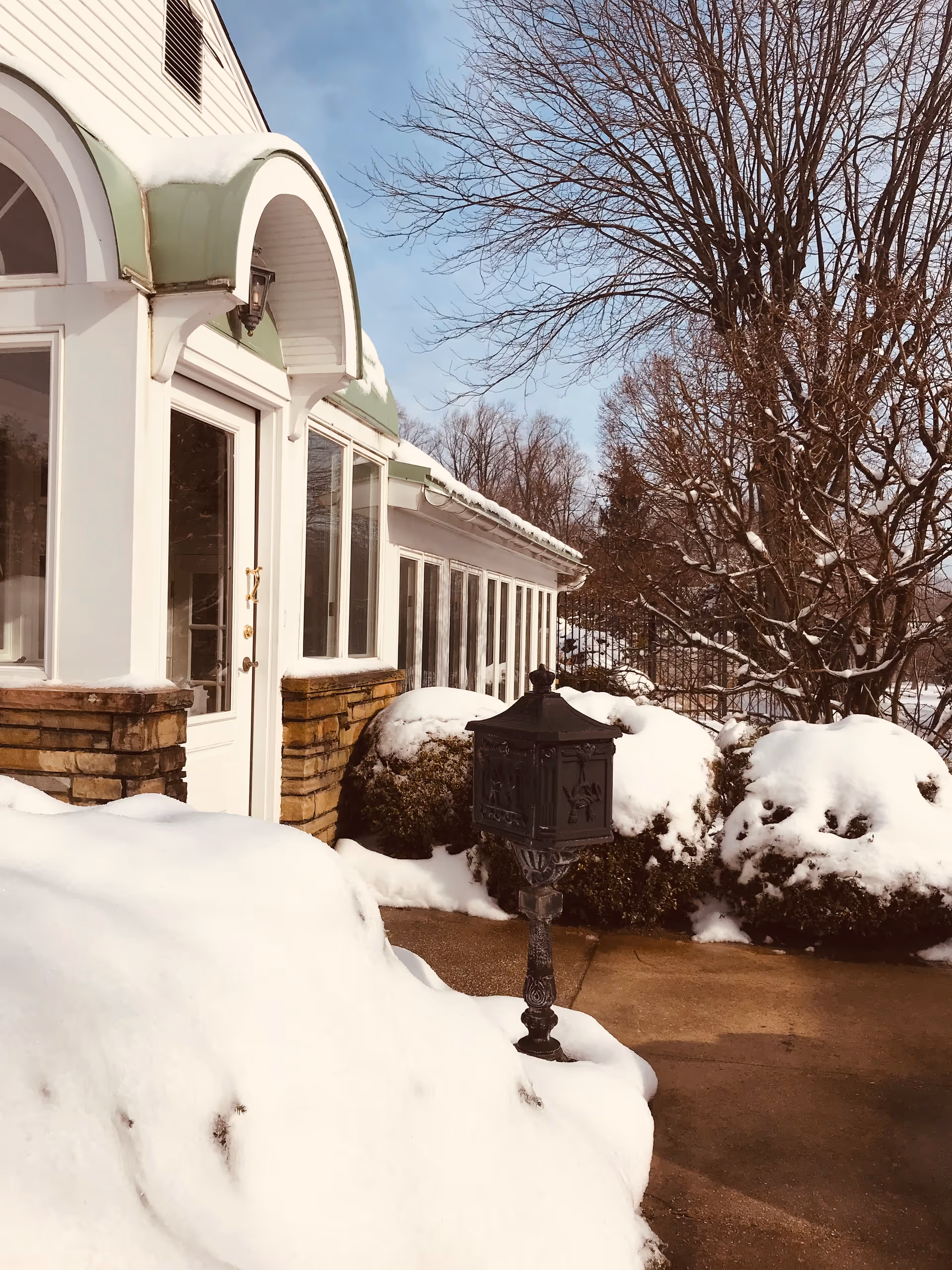 Snow covers the front of a facility with arched entry, lamp post, and bare trees.