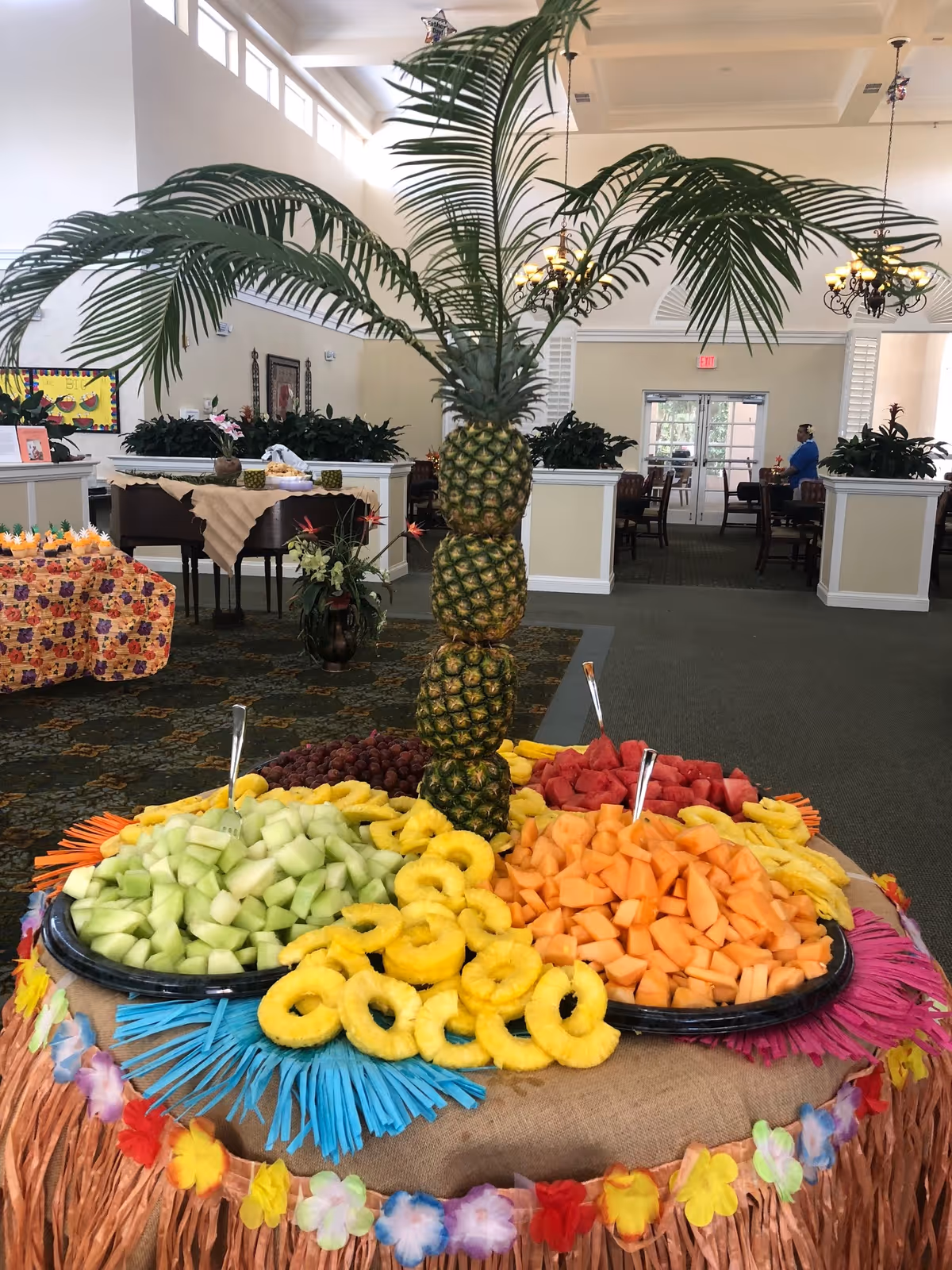 A festive fruit display in a senior living facility dining area featuring a centerpiece made of stacked pineapples with palm leaves on top, surrounded by trays of cut melon, watermelon, grapes, and pineapple rings on a table decorated with colorful fringe and flower garlands.