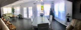 Sunlit communal room with a long dining table, upholstered chairs, sofas, and tall windows with sheer curtains.