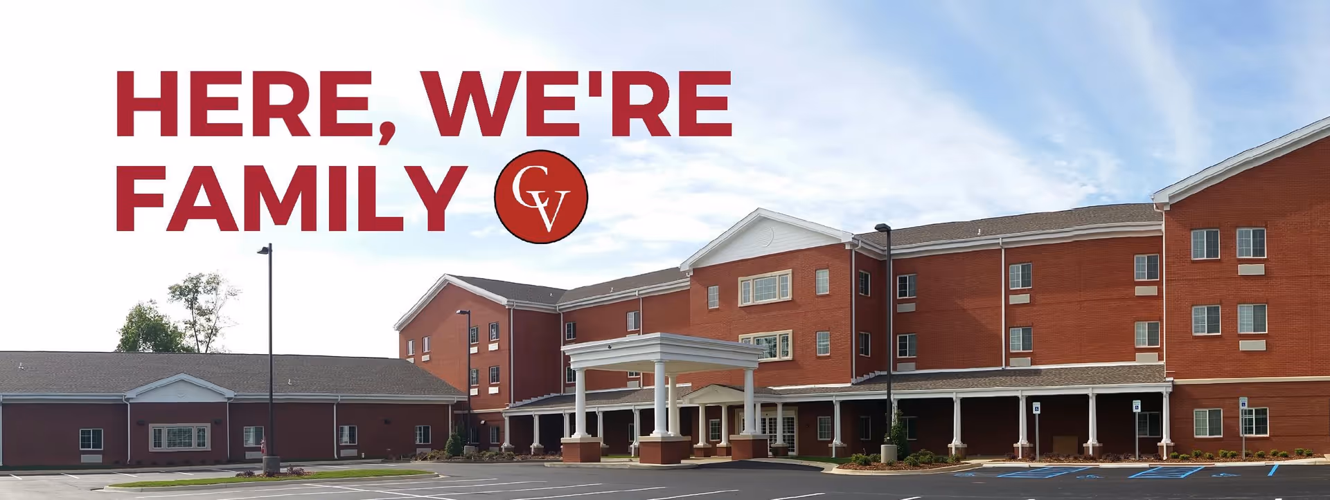 Exterior view of a large red brick senior living facility building with multiple windows and a covered entrance. The parking lot in front is mostly empty. Text on the image reads 'HERE, WE'RE FAMILY' with a circular logo containing the letters 'CV'.