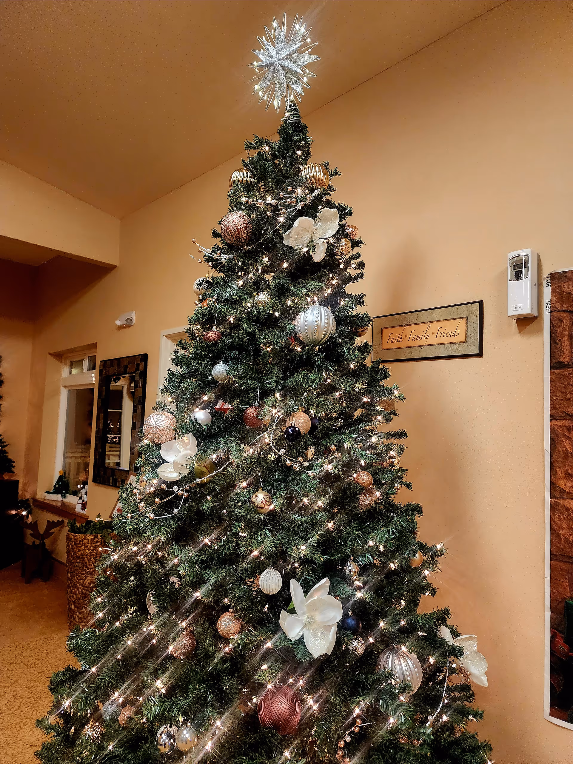 A decorated Christmas tree with white flowers, gold and silver ornaments, and string lights in a warmly lit room. A star topper shines brightly at the top of the tree. On the wall behind the tree is a framed sign that reads 'Faith Family Friends'.