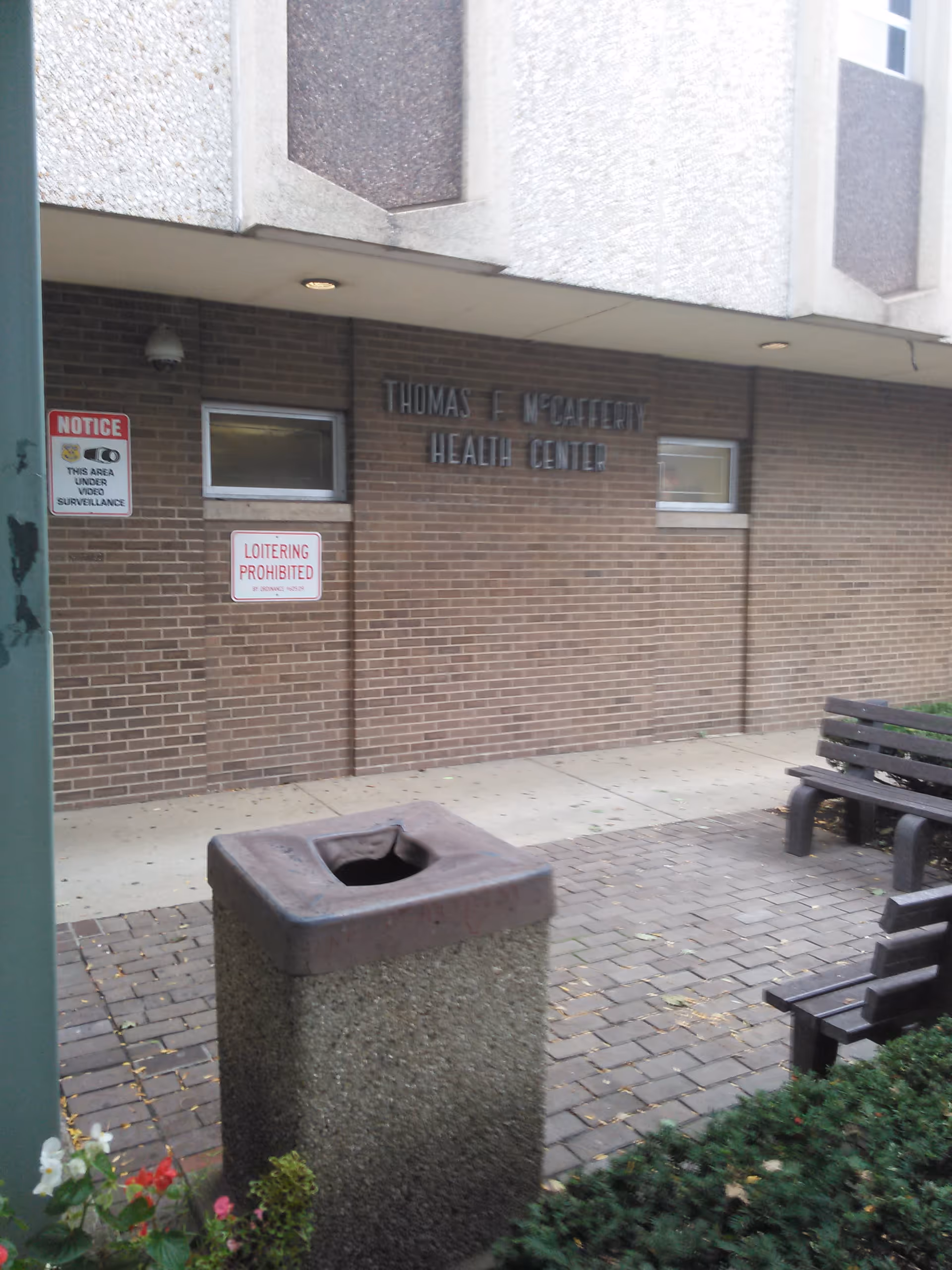 Front of Thomas F. McCafferty Health Center with brick wall, benches, and a trash can.