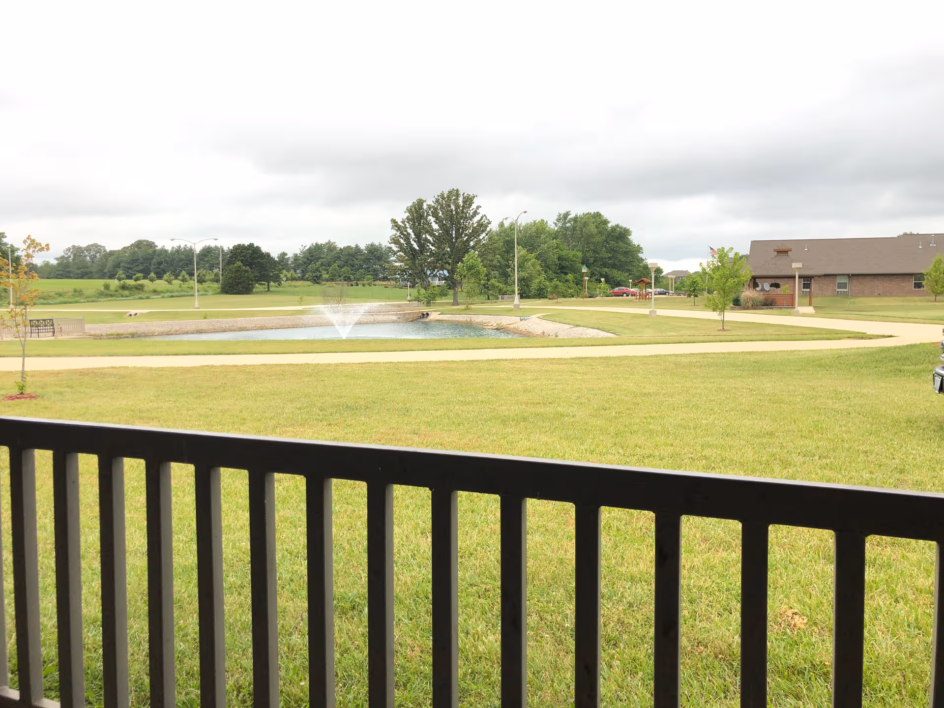 Balcony view overlooking green lawn, pond with fountain, walking paths, and distant facility building.