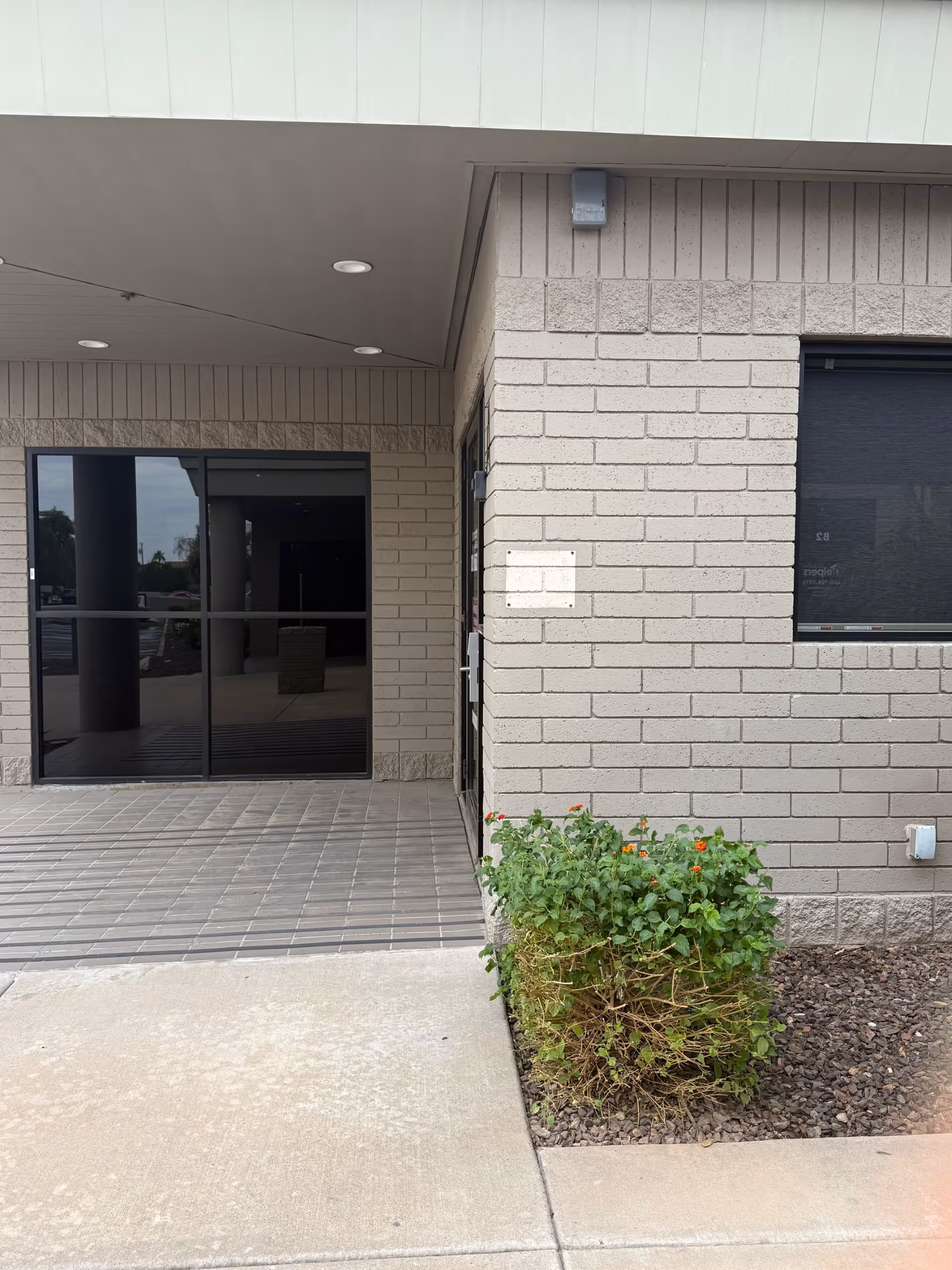 Exterior entrance of a beige brick facility with glass doors and a small plant bed.