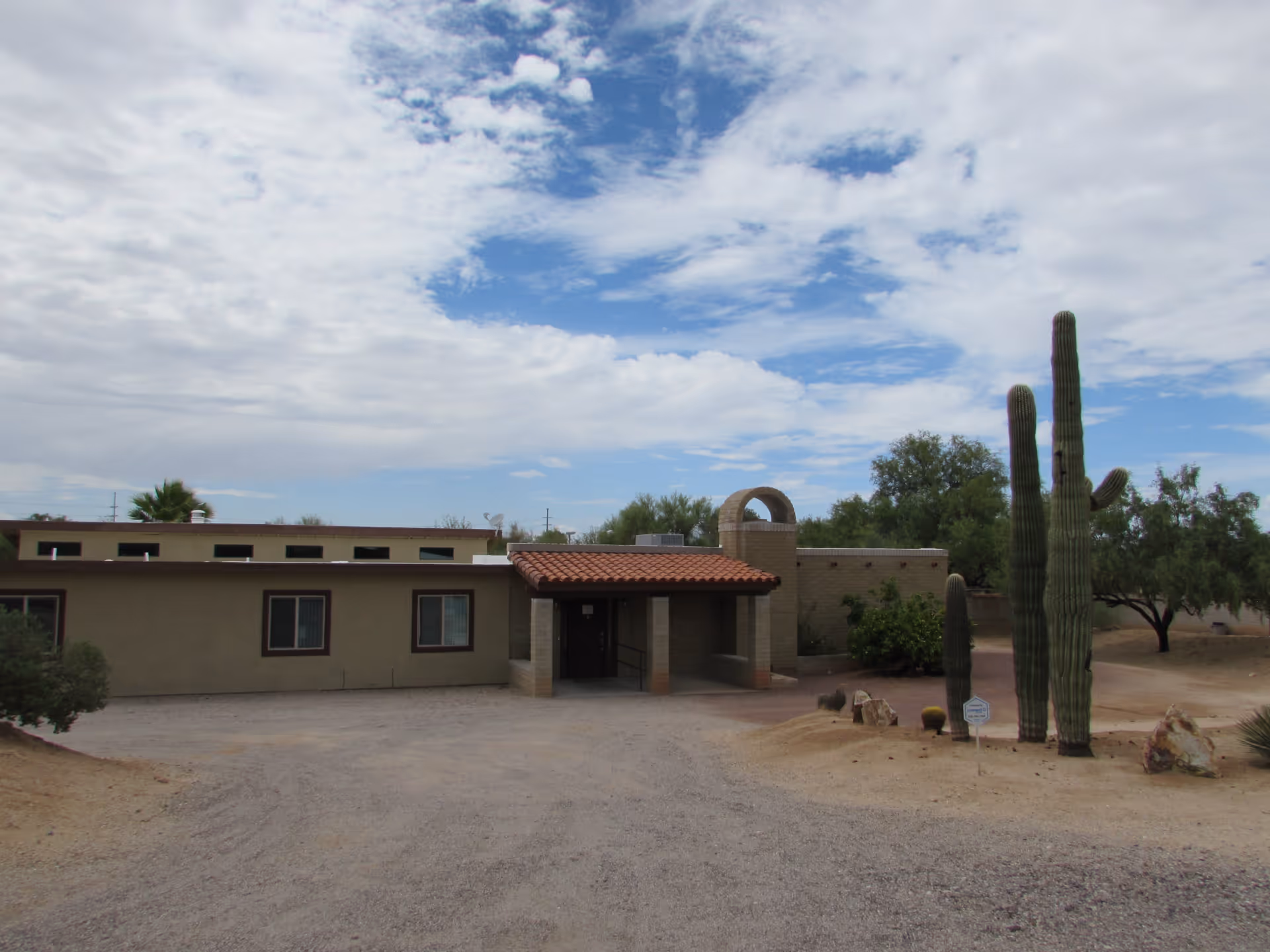 Single-story stucco building with a tiled entryway and tall saguaro cacti in a desert landscape under a partly cloudy sky.