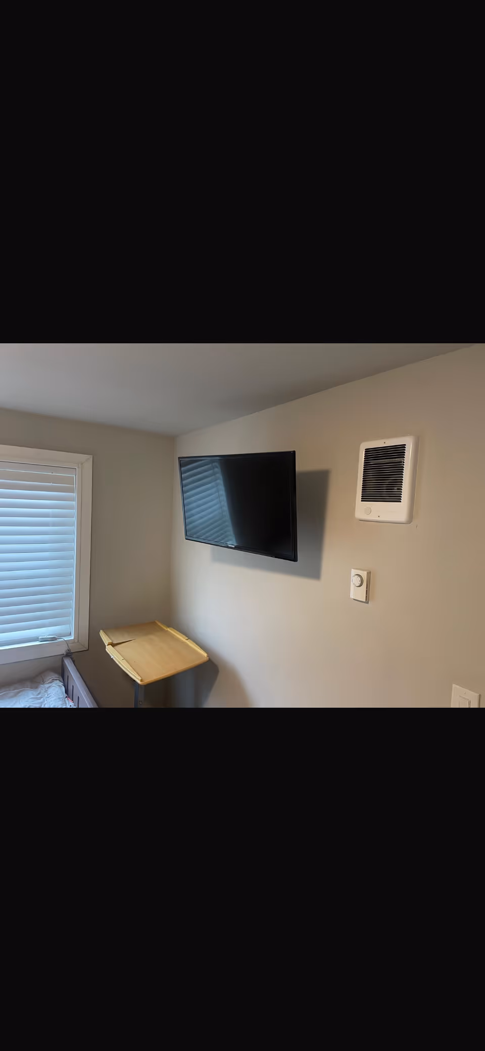 Bedroom with window blinds, wall-mounted TV, and a small wall-mounted desk.