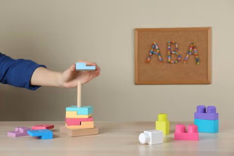 A hand stacks colorful wooden blocks on a table, with an ABA corkboard on the wall.