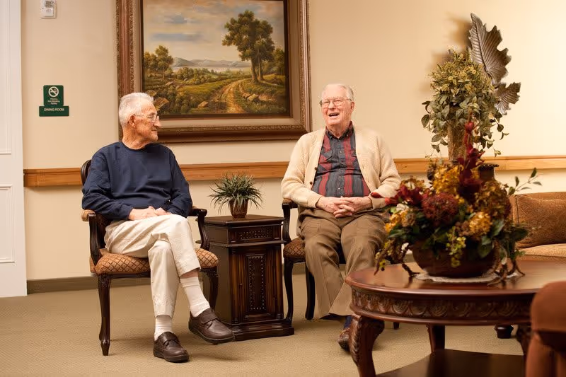 Two elderly men sitting and chatting in a furnished common lounge with chairs, a side table, framed landscape painting, and a floral centerpiece.