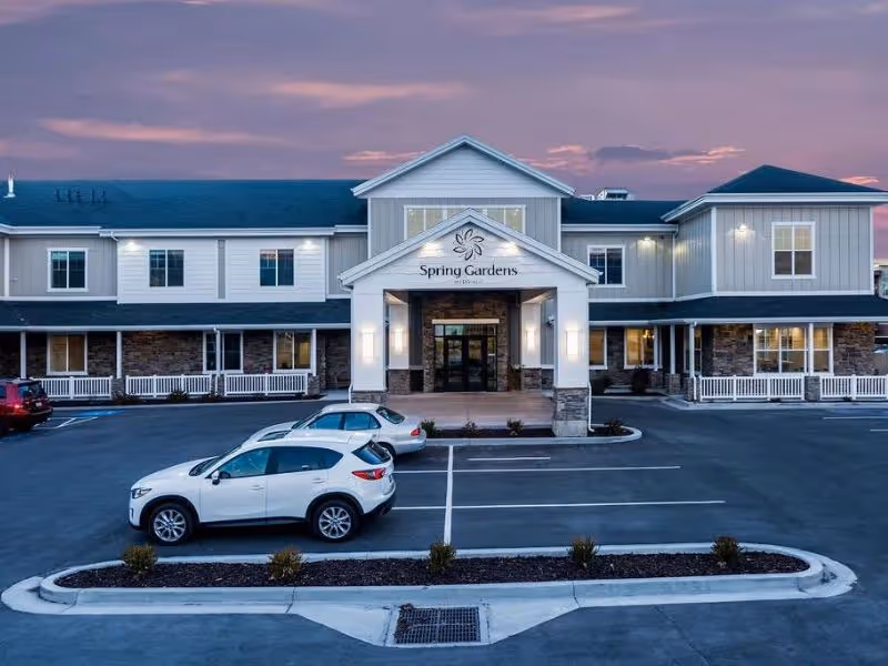 Front exterior of the Spring Gardens Senior Living Midvale building at dusk with a parking lot and several parked cars in front.